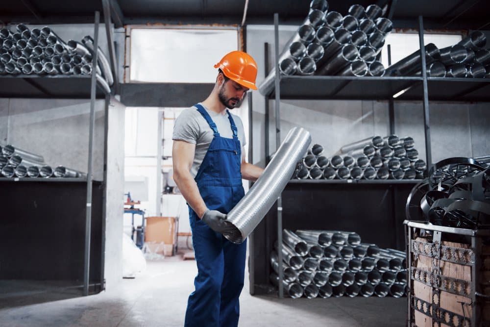 A worker in a hard hat carrying a length of metal ducting through a stores area